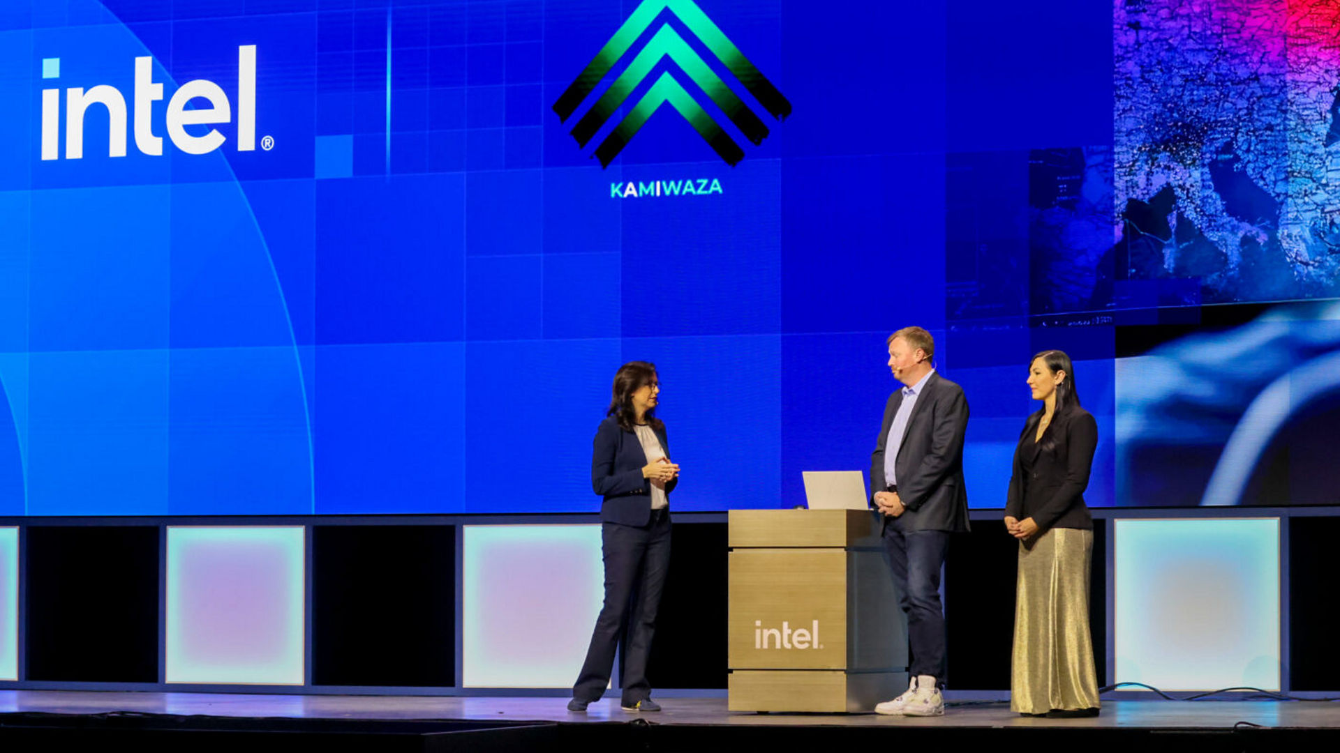 Three individuals stand on a stage in front of screens displaying the Intel logo and another logo labeled KAMIWAZA. One person is behind a podium with a laptop. The setting appears to be a professional or tech event.