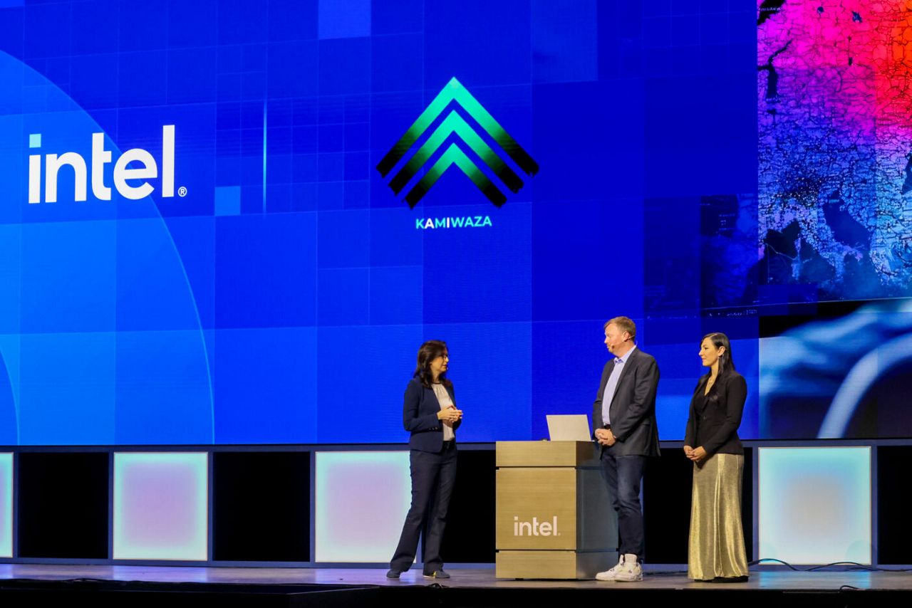 Three individuals stand on a stage in front of screens displaying the Intel logo and another logo labeled KAMIWAZA. One person is behind a podium with a laptop. The setting appears to be a professional or tech event.