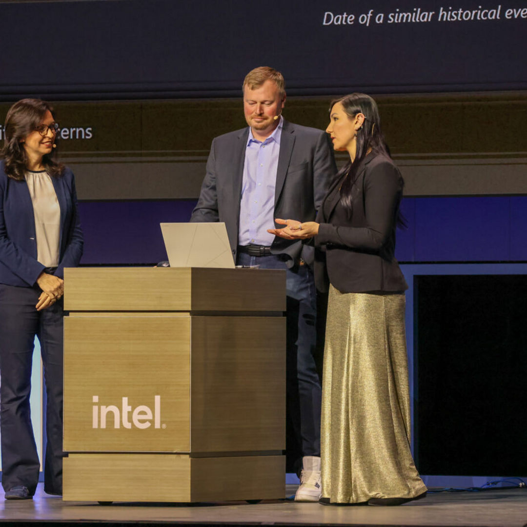 Three individuals are standing behind a podium labeled Intel on a stage. One person is speaking while the others listen. A laptop is open on the podium. The stage background is dark with partial text visible.