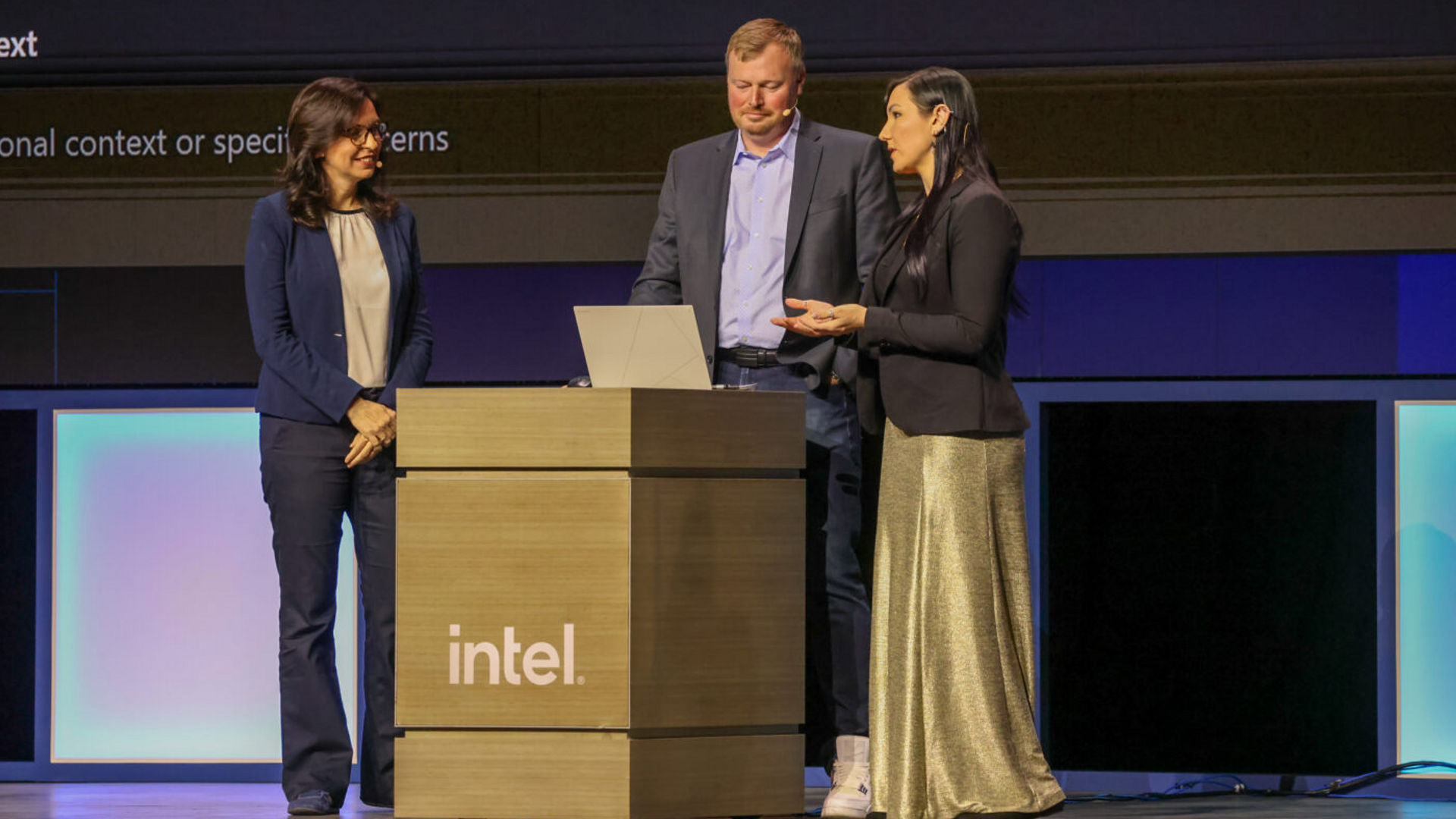 Three individuals are standing behind a podium labeled Intel on a stage. One person is speaking while the others listen. A laptop is open on the podium. The stage background is dark with partial text visible.