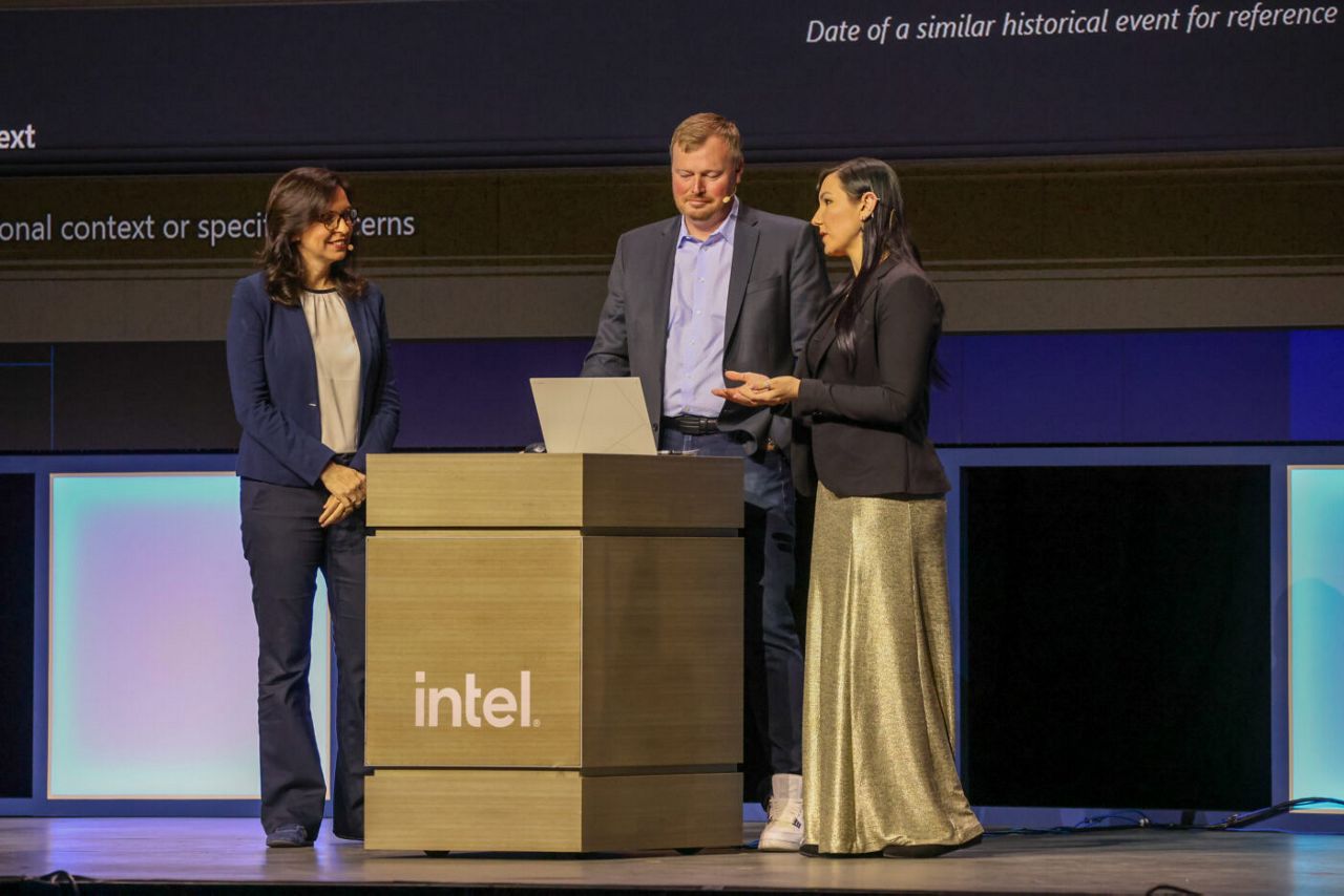 Three individuals are standing behind a podium labeled Intel on a stage. One person is speaking while the others listen. A laptop is open on the podium. The stage background is dark with partial text visible.