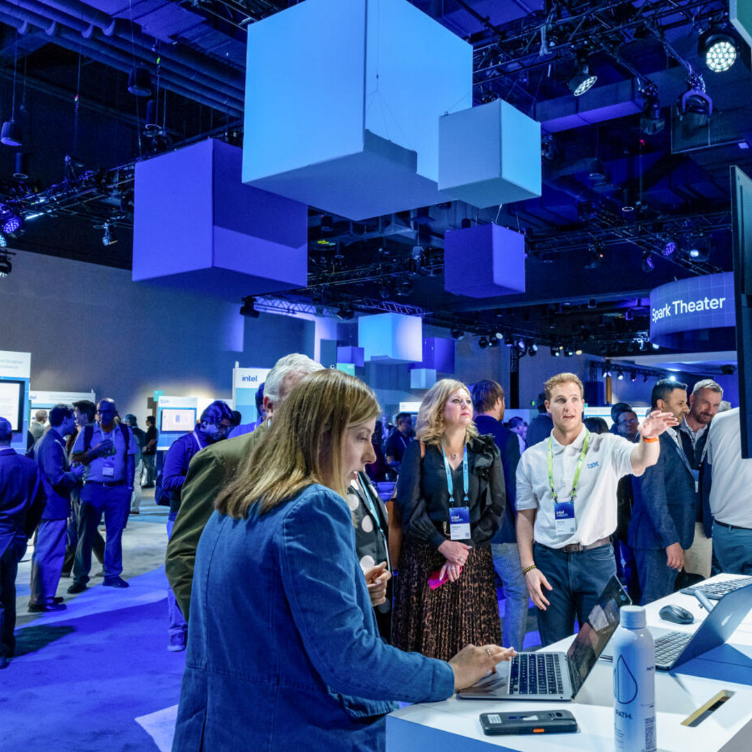 People are gathered at a tech conference, engaging with digital displays. A woman uses a laptop at a booth, while a man gestures towards a screen. Modern blue and white lighting and decor create a futuristic atmosphere.