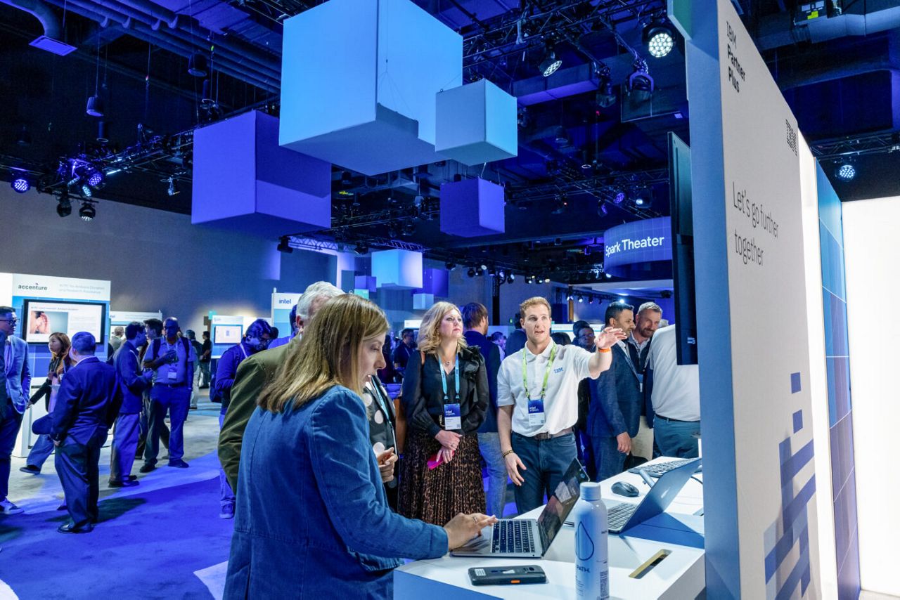 People are gathered at a tech conference, engaging with digital displays. A woman uses a laptop at a booth, while a man gestures towards a screen. Modern blue and white lighting and decor create a futuristic atmosphere.