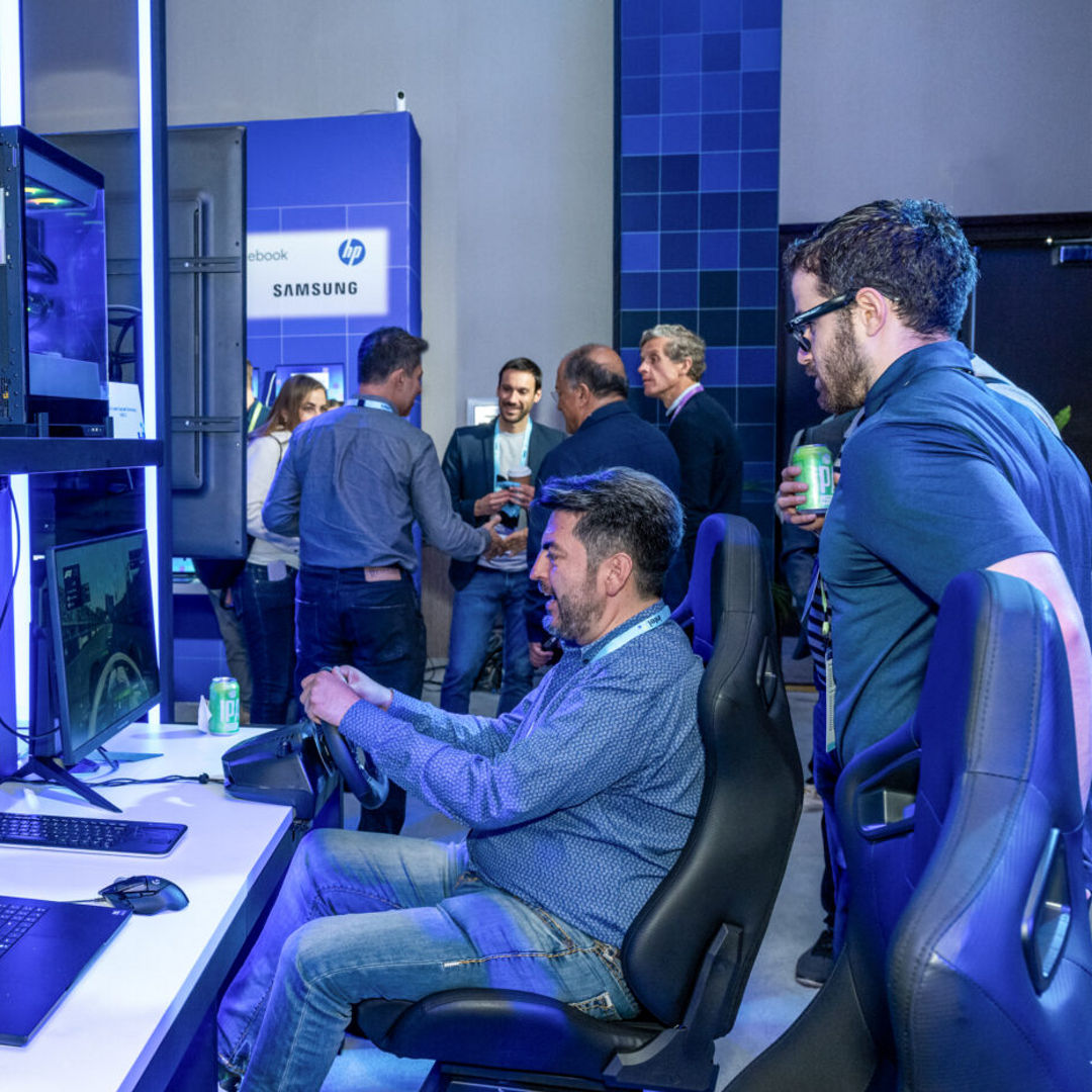 A group of people are gathered in a tech exhibit space. In the foreground, a man is seated at a racing simulation setup with multiple screens and a steering wheel. Others are standing and talking, with technology displays and brand logos in the background.