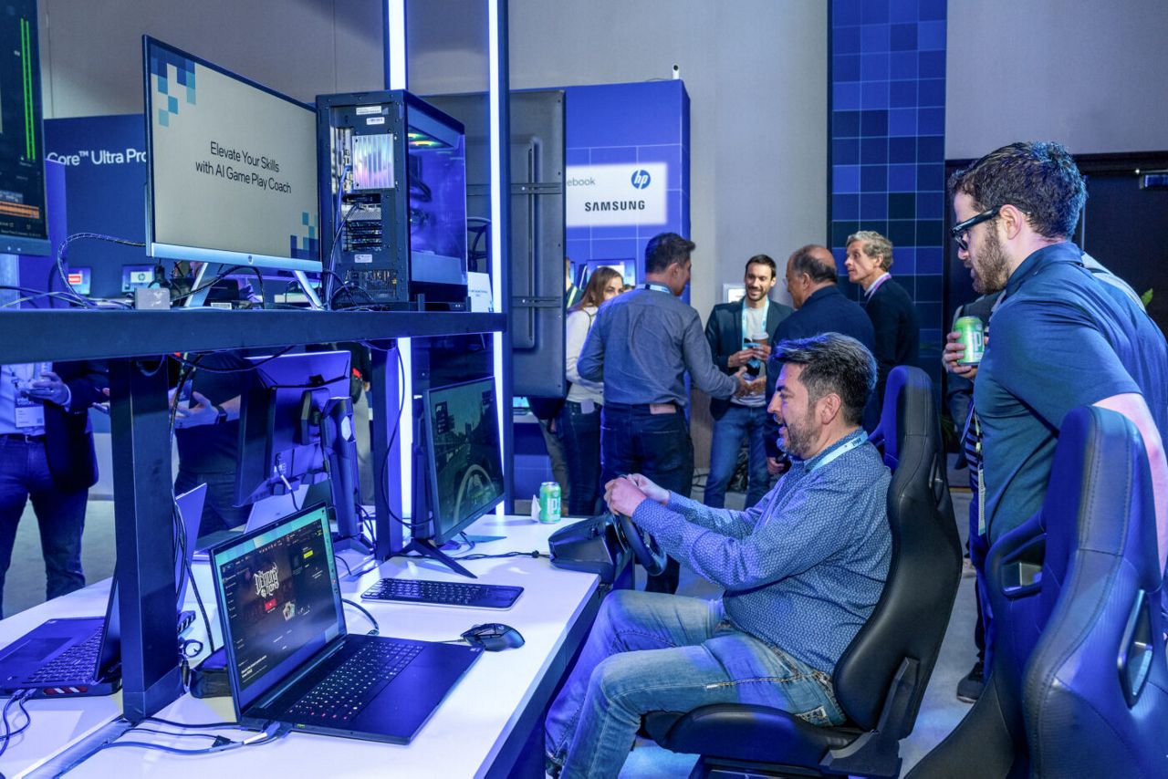A group of people are gathered in a tech exhibit space. In the foreground, a man is seated at a racing simulation setup with multiple screens and a steering wheel. Others are standing and talking, with technology displays and brand logos in the background.
