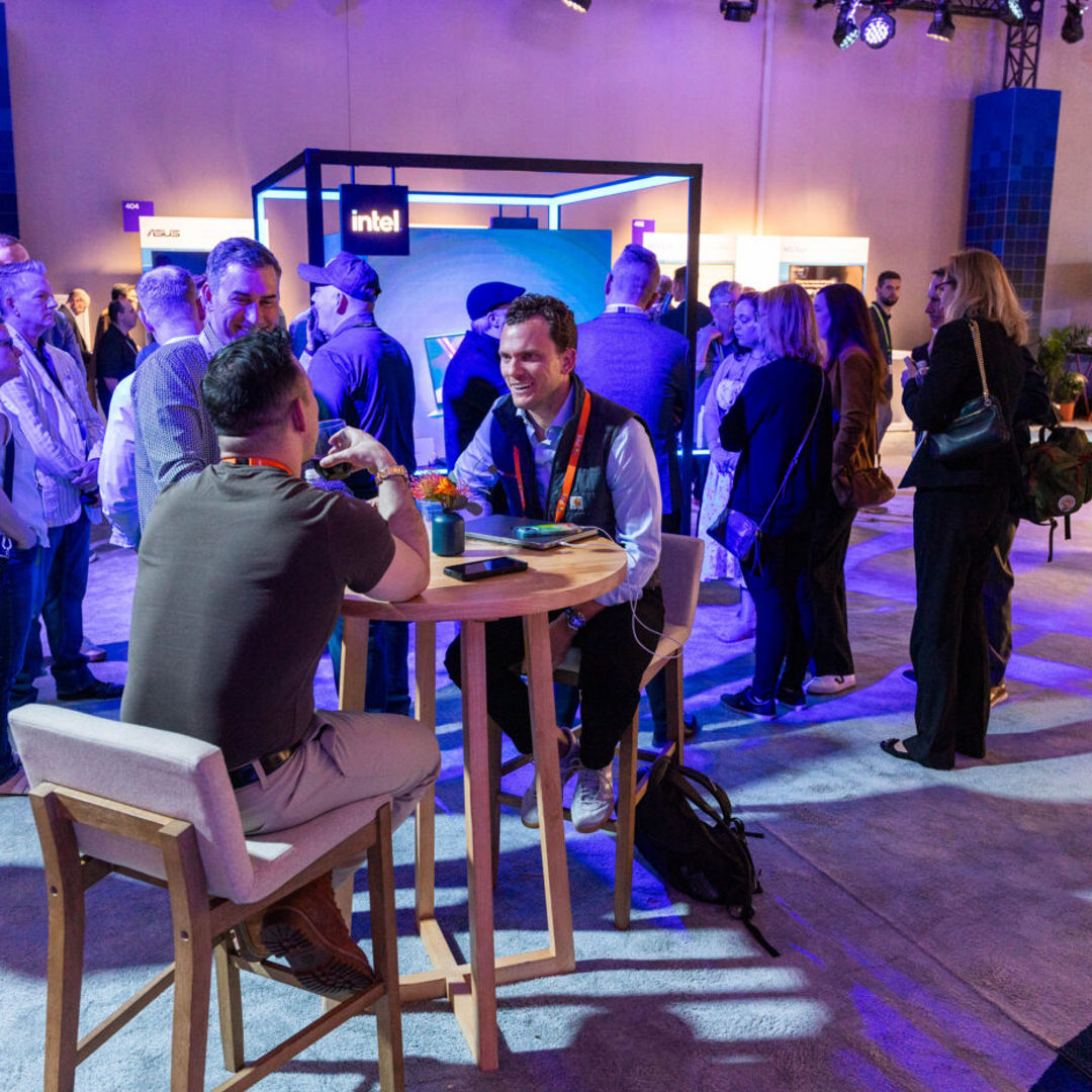 A group of people gathers at a tech expo. In the foreground, two men sit at a high table, engaged in conversation, while others stand and mingle in the background. Bright lighting and various tech company booths are visible.