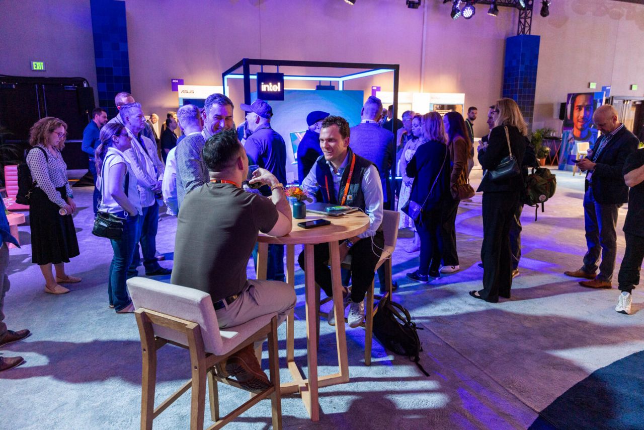 A group of people gathers at a tech expo. In the foreground, two men sit at a high table, engaged in conversation, while others stand and mingle in the background. Bright lighting and various tech company booths are visible.