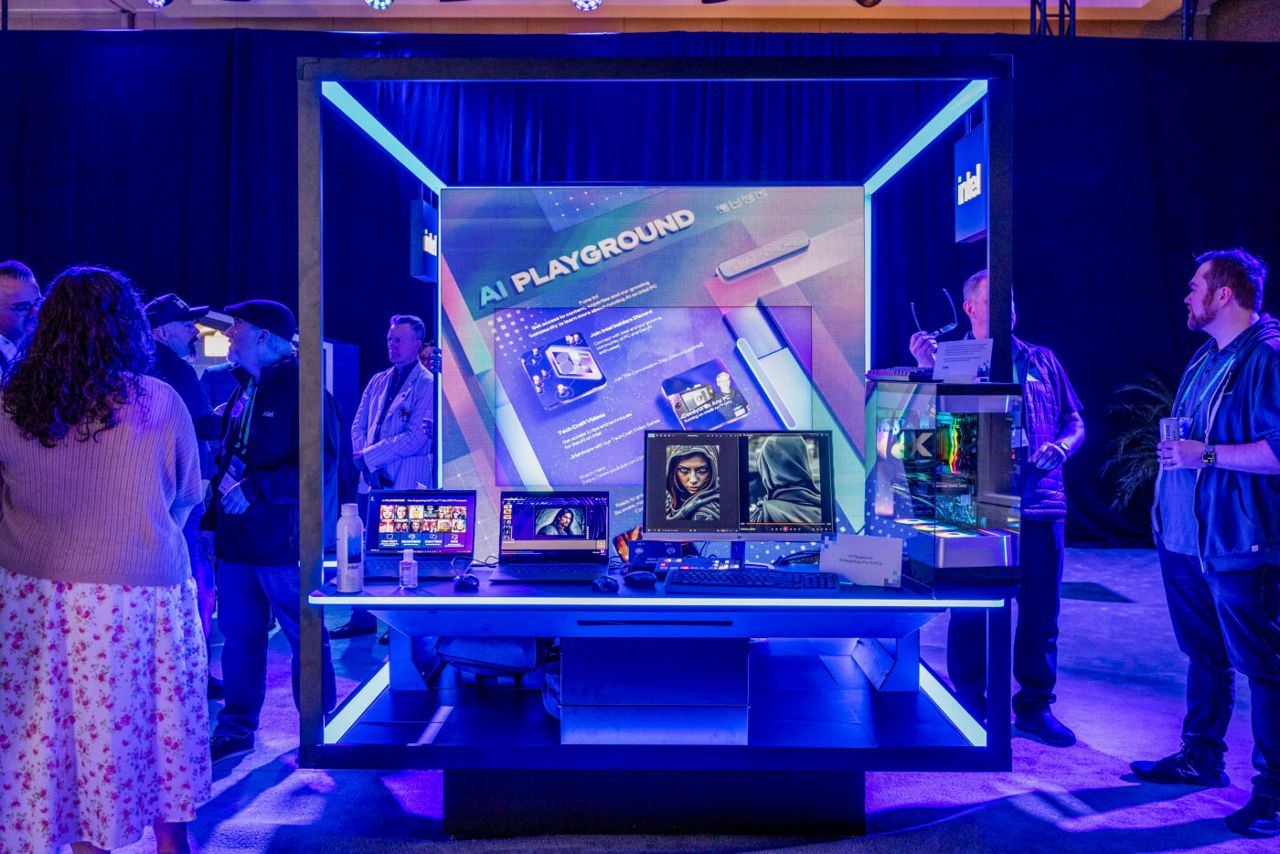 A tech exhibit labeled AI Playground features screens displaying digital graphics. Surrounded by blue lighting, attendees observe and interact with the setup. The atmosphere is vibrant and futuristic.