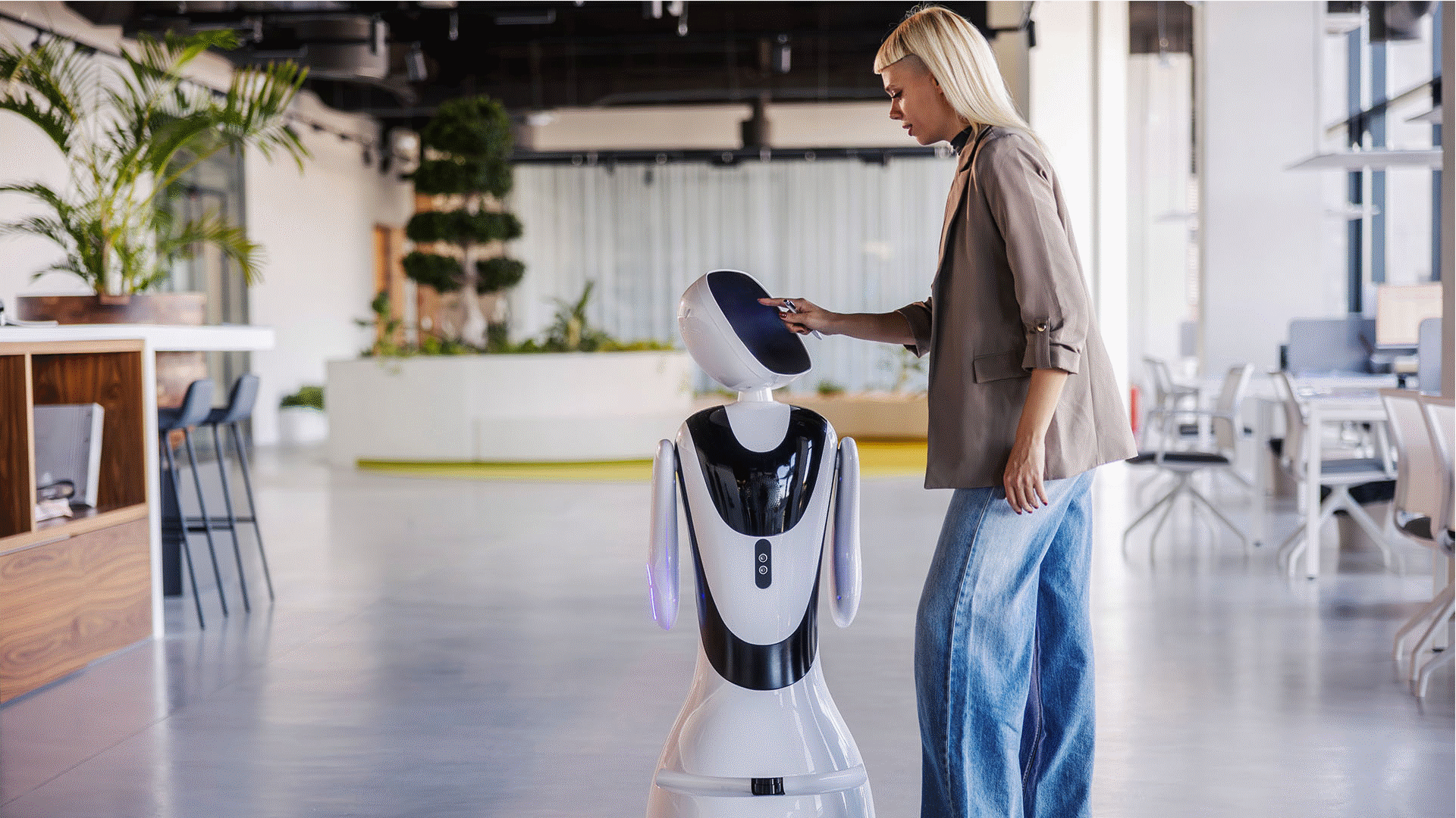 Young adult woman touching screen of service robot, exploring technology and artificial intelligence in a contemporary workplace