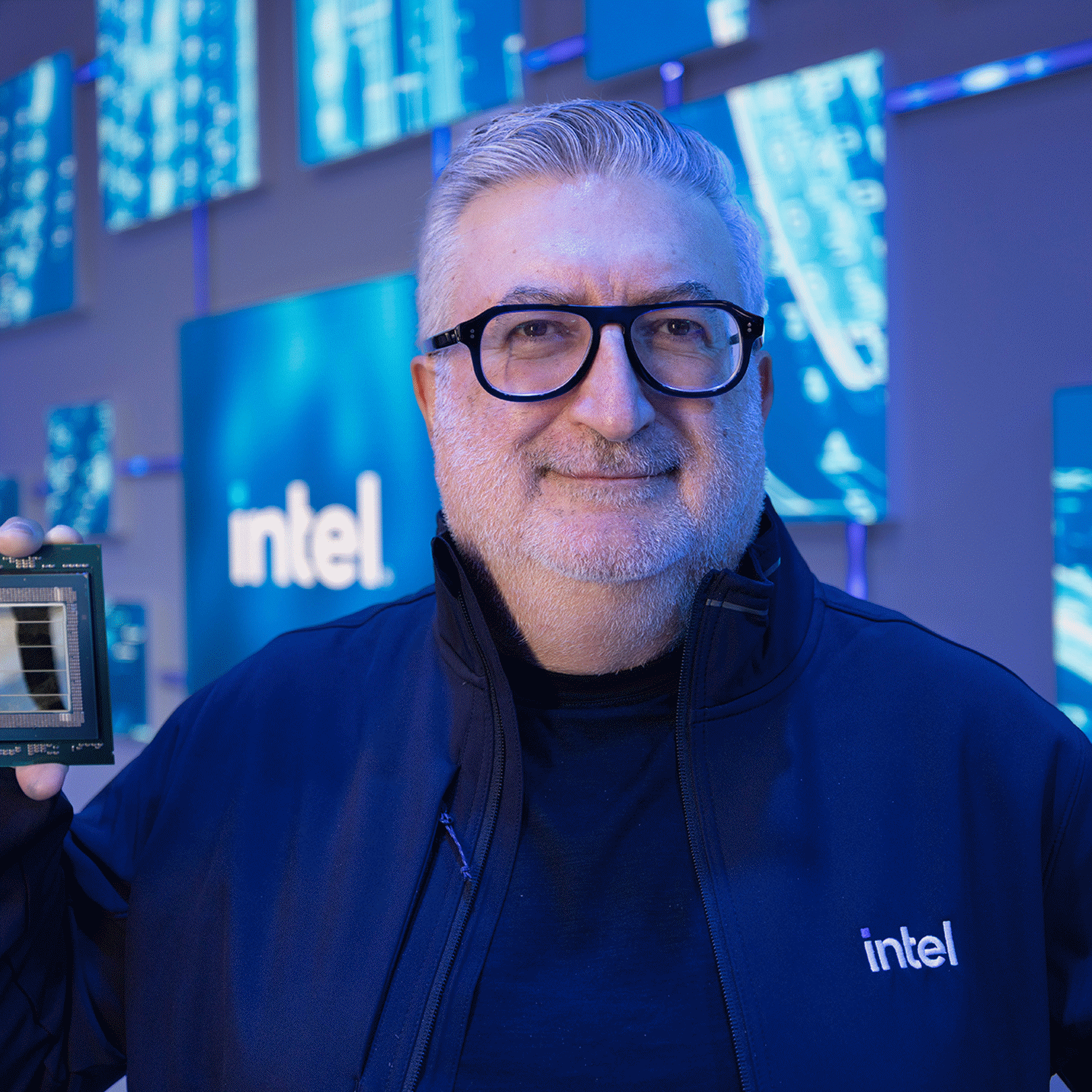 A man wearing an Intel jacket holds a processor in front of a blue Intel display.