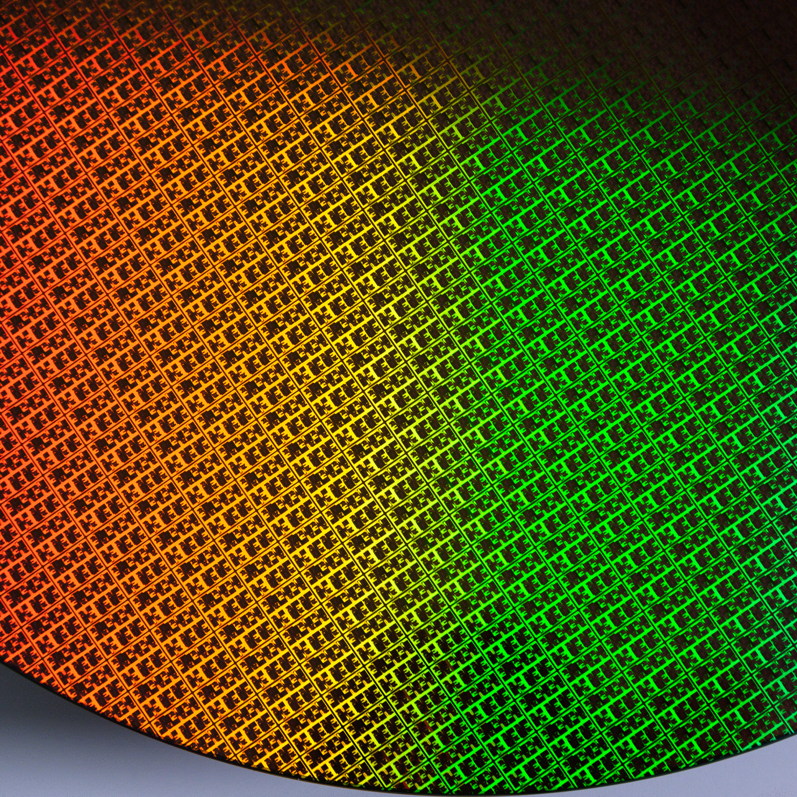 A close-up of a brightly colored silicon wafer, displaying a grid pattern. The surface reflects a spectrum of colors, blending from orange on the left to green on the right, showcasing the intricate and reflective nature of the wafer.
