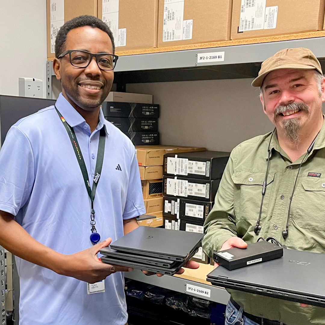 Two smiling men in a storage room hold laptops. One wears a light blue polo and glasses, the other a green shirt and hat. Shelves behind them are filled with boxes and more laptops.
