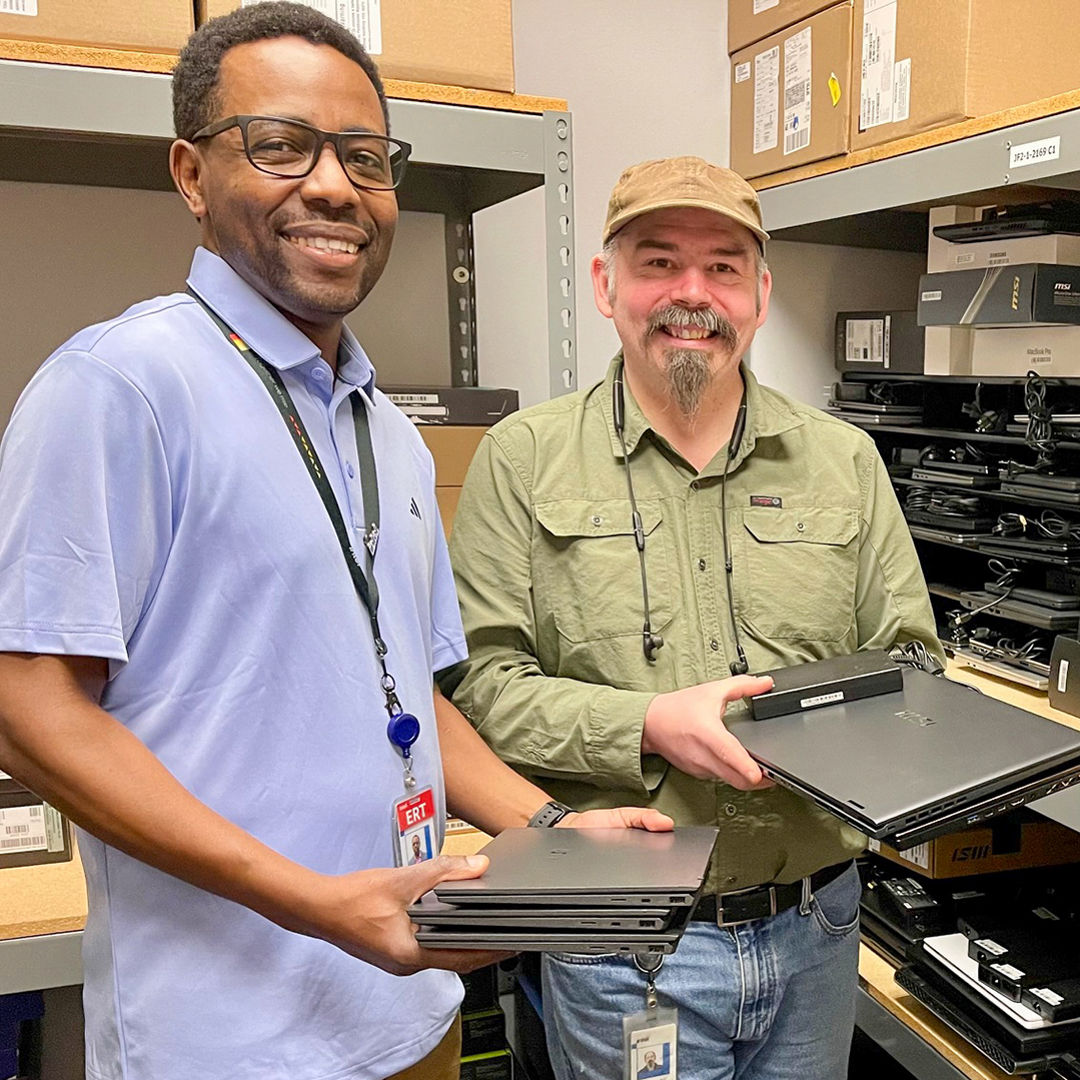 Two men smiling while holding laptops in a storage room. Shelves around them are stocked with computer equipment, including boxes labeled Mac Studio. One man is wearing a light blue shirt, the other in a green shirt and cap.
