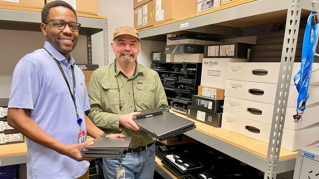 Two men smiling while holding laptops in a storage room. Shelves around them are stocked with computer equipment, including boxes labeled Mac Studio. One man is wearing a light blue shirt, the other in a green shirt and cap.