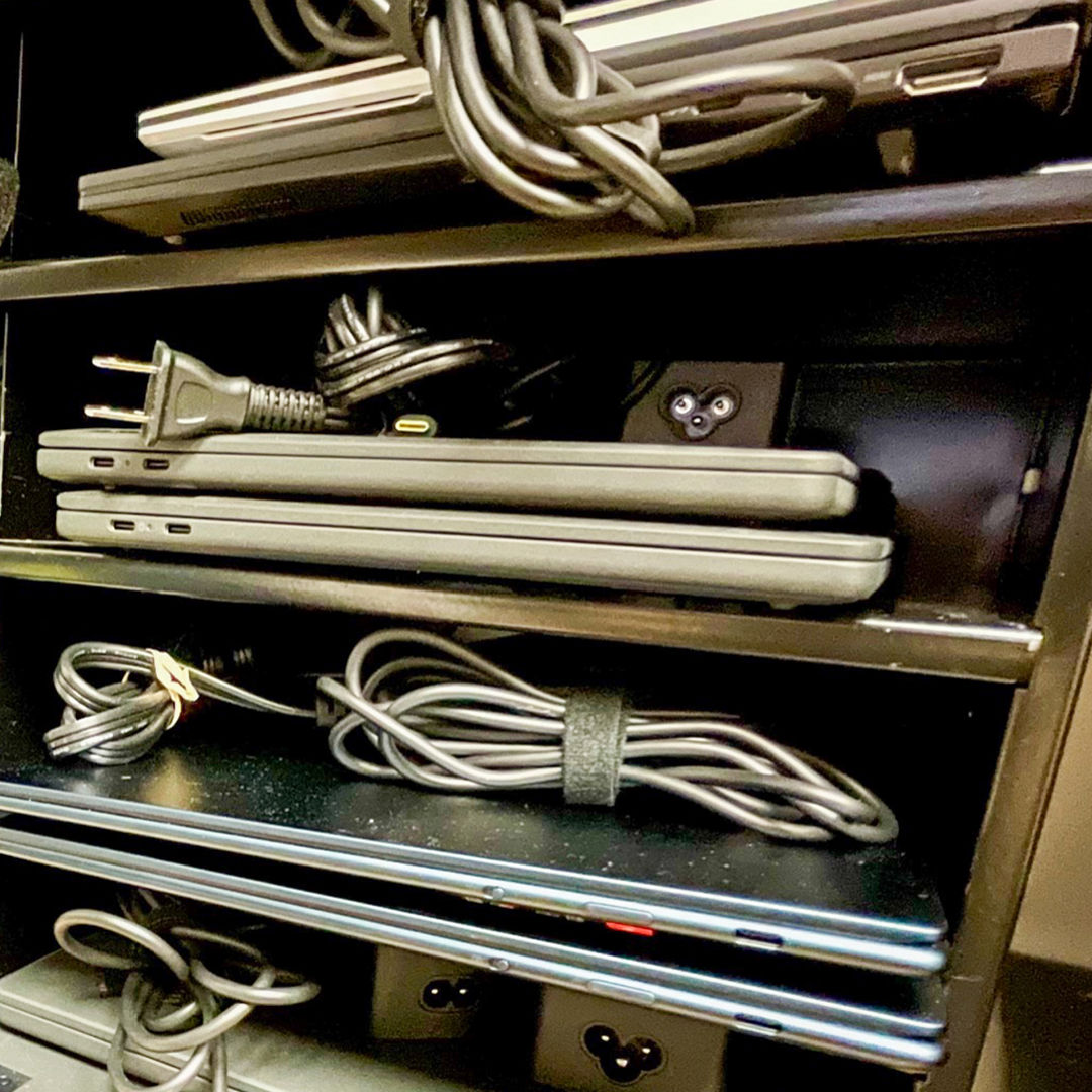 Shelves filled with stacked laptops, each with its power cable coiled neatly. The shelves contain a mix of closed laptops and power adapters, organized in rows.