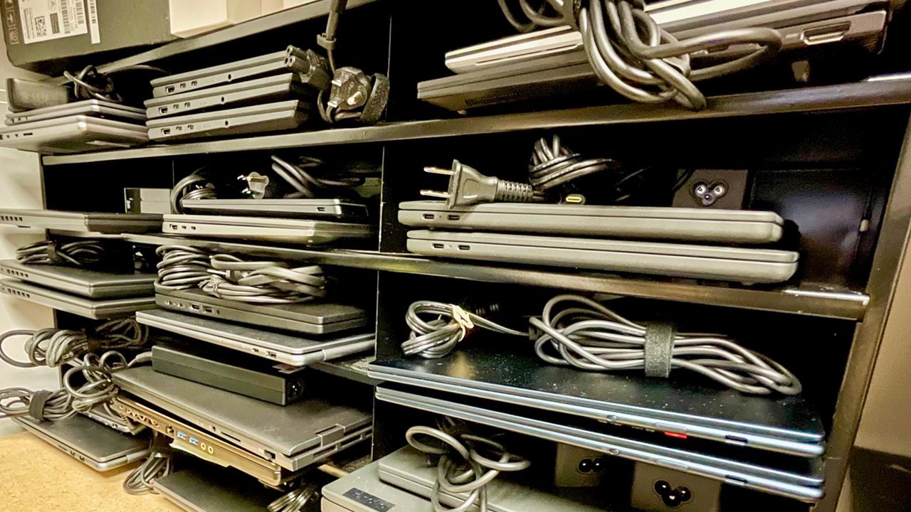 Shelves filled with stacked laptops, each with its power cable coiled neatly. The shelves contain a mix of closed laptops and power adapters, organized in rows.