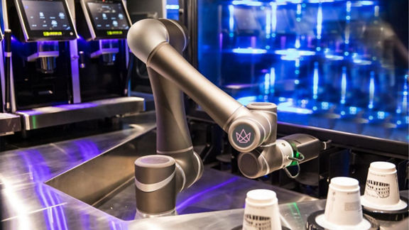 Roboterarm auf einem Edelstahltresen in einem Café, mit drei Reihen von To-Go-Tassen, die in Reichweite des Arms umgekehrt gestapelt sind