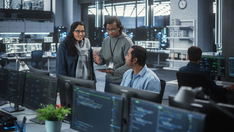 Three colleagues have a discussion in a modern work environment. One person sits at a workstation while the other two people stand in close proximity. One standing person holds a tablet