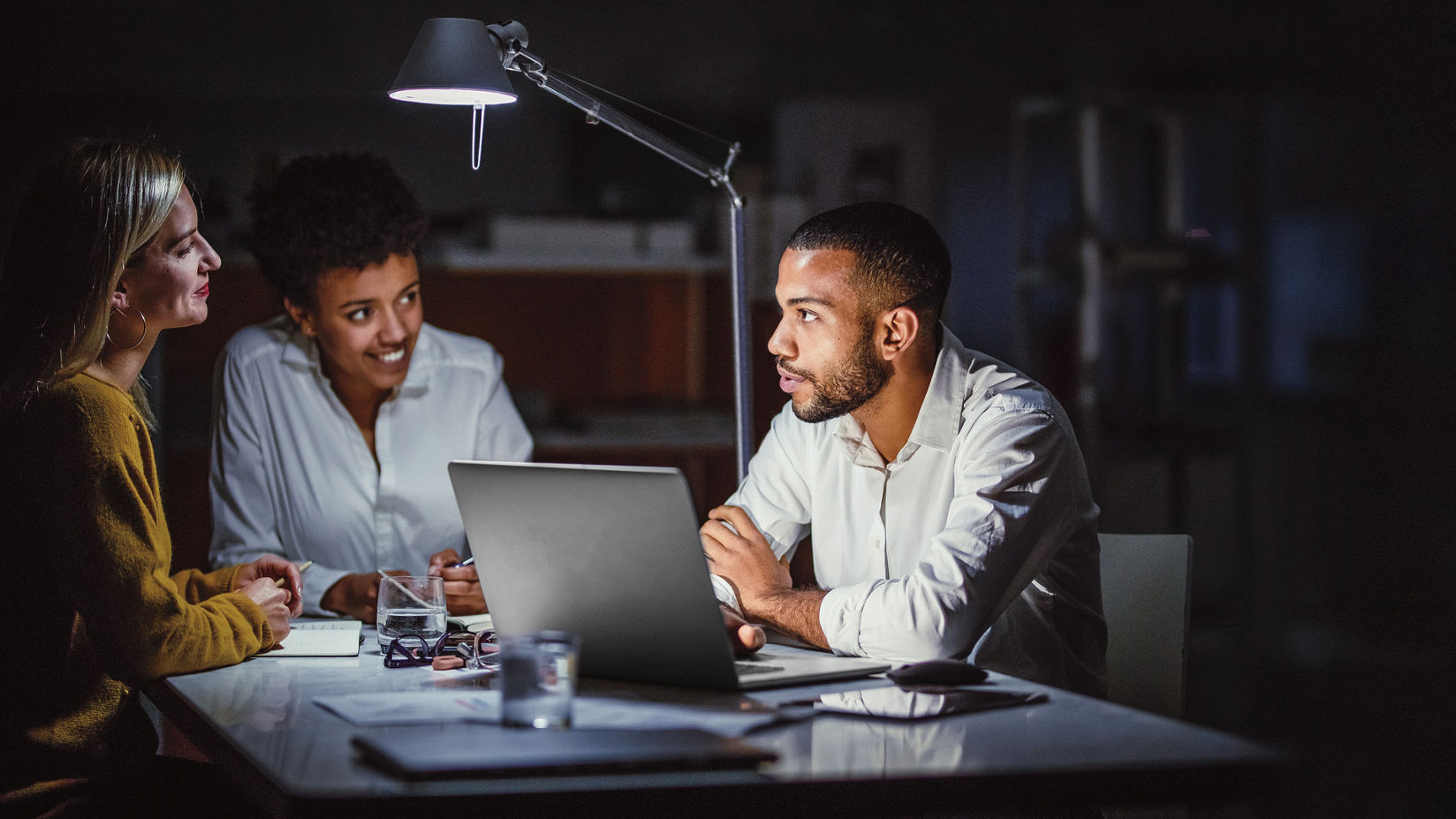 Tres compañeros de trabajo de empresa en un entorno de trabajo sentados en torno a una mesa colaborando. Una persona apoyando la mano en el teclado de una laptop