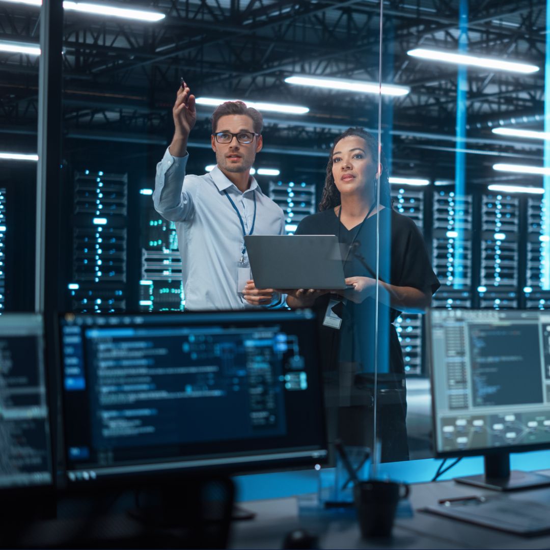 Two colleagues are in a data center. One is holding a laptop, and the other is pointing at something in the distance.